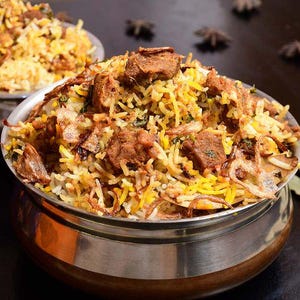 Puede incluir: Primer plano de un cuenco lleno de biryani, un plato de arroz mixto del sur de Asia. El plato contiene arroz amarillo, trozos de carne y cebollas fritas crujientes. El cuenco es de acero inoxidable con una base de madera.