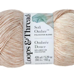 May include: A skein of Soft Ombre polyester blend yarn in a neutral color palette. The yarn is a mix of light beige and peach tones, with a label that reads "Soft Ombre" and "Ombrée Douce". The skein contains 426 yards or 5.29 ounces of yarn.