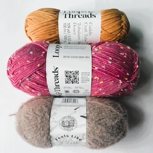 May include: Three skeins of yarn in shades of orange, pink, and taupe. The top skein is orange with a white label that reads "Loops & Threads." The middle skein is pink with colorful flecks and a white label. The bottom skein is taupe with a "Lion Brand Yarns" label.