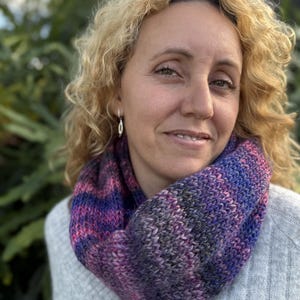 May include: A hand-knitted infinity scarf featuring a variegated yarn pattern in shades of deep purple, magenta, and charcoal gray. The scarf is crafted with a chunky knit texture and is worn around the neck over a light gray textured sweater.