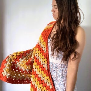 May include: A handmade crochet shawl featuring a vibrant granny square pattern in alternating stripes of bright orange, golden yellow, and cream yarn. The textile accessory has a textured, open-weave construction with a solid orange border edge.