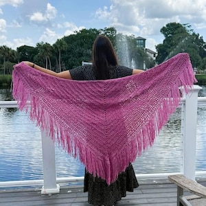 May include: A triangular crochet shawl in a vibrant magenta hue, featuring a textured stitch pattern and long, flowing fringe along the bottom edges. The garment is displayed spread out to showcase its wide, pointed shape and intricate yarn work.