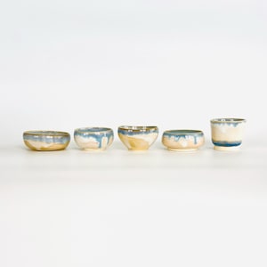 以下が含まれることがあります： 独特な釉薬を施した5つのセラミックボウルのセット。ボウルはサイズと形状が異なり、淡いブルーとベージュの配色に光沢のある仕上がりです。釉薬が側面を流れ落ち、手作りの美しさを添えています。