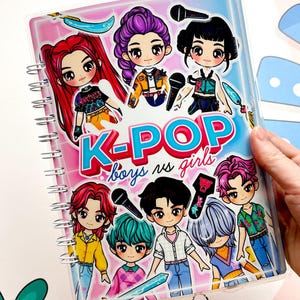 Può includere: Quaderno a spirale con personaggi dei cartoni animati K-POP. La copertina mostra il testo "K-POP boys vs girls" in un design vivace. Il quaderno ha una copertina di plastica trasparente.