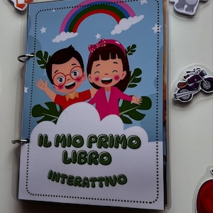 May include: An interactive children's book with a blue cover, a rainbow, clouds, and illustrations of two smiling children. The book's title, "IL MIO PRIMO LIBRO INTERATTIVO," is in white text. Animal stickers, including a duck and frog, surround the book.