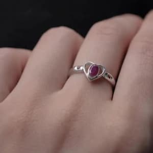 Peut inclure: Une bague en argent dotée d'un sertissage en forme de cœur encadrant une petite pierre précieuse rouge foncé de taille ovale. L'anneau est lisse et poli, avec le motif de cœur centré sur le doigt, présentant une finition métallique nette.