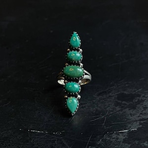 May include: A silver ring featuring five vertically aligned, oval-shaped turquoise stones. The stones are set in serrated silver bezels, with the centre stone being the largest, flanked by two smaller oval stones and two teardrop-shaped stones at the ends.
