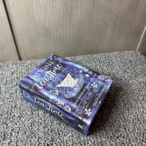 Puede incluir: Una caja rectangular titulada "Divine Tea Time Inspiration Cards" con un dise&ntilde;o azul oscuro y morado. La caja presenta las palabras "Tea Time" en una fuente cursiva, junto con ilustraciones florales y celestiales. Tambi&eacute;n es visible el nombre de la autora, Tracy Loughlin.