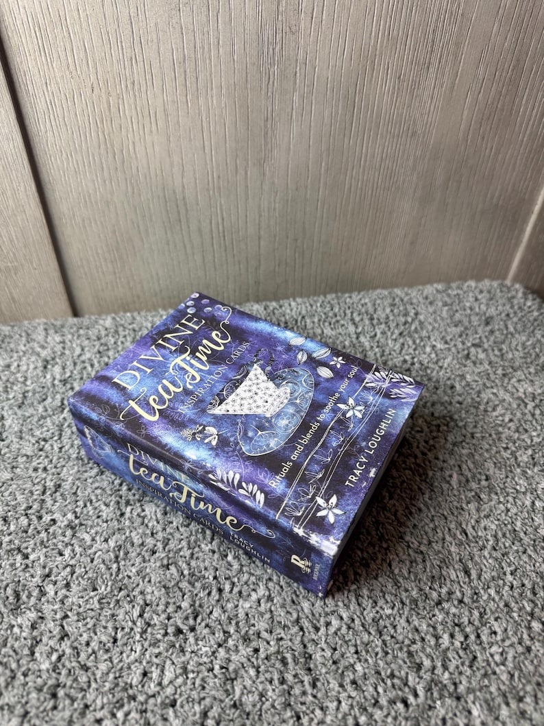 Puede incluir: Una caja rectangular titulada "Divine Tea Time Inspiration Cards" con un dise&ntilde;o azul oscuro y morado. La caja presenta las palabras "Tea Time" en una fuente cursiva, junto con ilustraciones florales y celestiales. Tambi&eacute;n es visible el nombre de la autora, Tracy Loughlin.