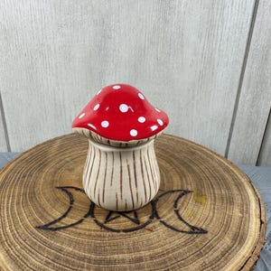 May include: A ceramic mushroom-shaped container with a red cap and white polka dots. The container's base is beige with vertical brown stripes. It sits on a wooden slice with a crescent moon design.