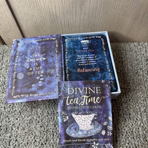 Puede incluir: Un juego de cartas de inspiraci&oacute;n "Divine Tea Time" con un dise&ntilde;o floral azul y morado. La caja incluye tarjetas con texto como "La respuesta es una taza de t&eacute;" y una receta de "T&eacute; de pimienta".