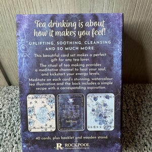 Puede incluir: Un juego de cartas con tem&aacute;tica de t&eacute; en tonos morados y azules, con el texto "Tea drinking is about how it makes you feel!" y "Uplifting, Soothing, Cleansing and so much more." El set incluye 40 cartas, un folleto y un soporte de madera.
