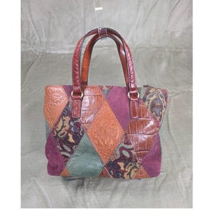 May include: A patchwork tote bag with a variety of textures and colors. The bag features diamond-shaped panels of leather, suede, and patterned fabric in shades of brown, green, and purple. It has two brown leather handles.