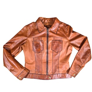 May include: A brown leather jacket with a collared neckline, two front pockets, and a zipper closure. The jacket has long sleeves with buttoned cuffs and a fitted waist. The leather has a distressed appearance with variations in color.