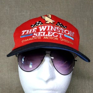 May include: A red baseball cap with a black brim featuring embroidered text that reads THE WINSTON SELECT and CHARLOTTE MOTOR SPEEDWAY, along with a gold eagle and two checkered flags. The cap is displayed on a white mannequin head with sunglasses.