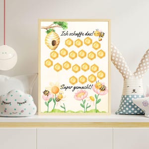 Op de afbeelding: Een ingelijste aquarelprint met een bijenkorf, bijen en bloemen. De print heeft de tekst "Ich schaffe das!" en "Super gemacht!" in een grillig ontwerp. De print bevat 20 gele honingraatvormen.
