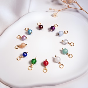 Può includere: Una collezione di ciondoli con perline colorate con anelli dorati. Le perline sono di vari colori, tra cui blu, viola, rosso, verde e bianco. I ciondoli sono disposti su una superficie bianca.