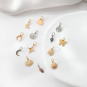 Op de afbeelding: Een verzameling kleine, decoratieve bedels in diverse vormen, waaronder een vlinder, hart, halve maan, ster en bloem. De bedels zijn in goud-, zilver- en bronskleuren, sommige met ingewikkelde ontwerpen. Ze worden gepresenteerd op een witte ondergrond.