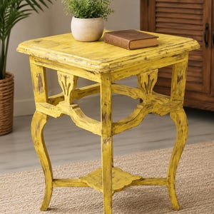 May include: A distressed yellow side table with a square top and curved legs. A small potted plant and a brown book rest on the table. The table has decorative cutouts and a lower shelf. The table is approximately 24 inches tall.