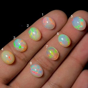 Könnte beinhalten: Acht oval geformte Cabochon-Opale, die mit den Zahlen eins bis acht beschriftet sind. Jeder Edelstein zeigt eine milchig-weiße Basis mit irisierenden Farbspielen in Grün, Orange und Pink. Die Steine sind in einer Sequenz angeordnet.