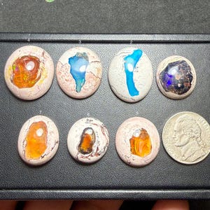 Puede incluir: Una colección de piedras preciosas de ópalo ovaladas en varios colores, incluyendo naranja, azul y púrpura, sobre un fondo neutro. Se incluye una moneda para comparar el tamaño. Los ópalos tienen patrones internos únicos.
