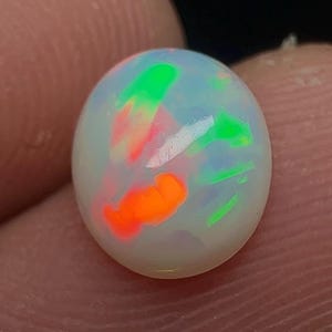 Puede incluir: Una piedra preciosa de ópalo blanco de forma ovalada y lisa que presenta destellos vibrantes de color naranja neón y verde brillante en su superficie pulida. La piedra muestra una base lechosa translúcida con manchas de color.