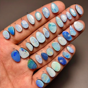 Pode incluir: Uma coleção de trinta e cinco opalas polidas, de formato oval e gota, em vários tons de azul, branco e verde iridescente. Cada pedra preciosa é numerada individualmente de um a trinta e cinco, exibindo jogos de cores e padrões únicos.