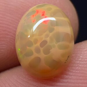 May include: An oval-shaped cabochon opal gemstone featuring a translucent, pale peach-coloured body with a distinct honeycomb pattern. The stone displays vibrant flashes of bright red and subtle green play-of-colour across its smooth, polished surface.
