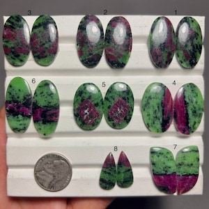 May include: A collection of polished zoisite and ruby cabochons in various oval and teardrop shapes, featuring green, black, and magenta patterns. The stones are arranged in rows on a white display tray, accompanied by a US nickel for scale.