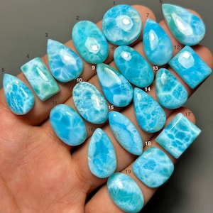 May include: A collection of nineteen polished Larimar gemstones in various shapes including teardrop, oval, and rectangular forms. Each stone displays a vibrant sky-blue color with white, cloud-like patterns and a smooth, reflective surface finish.
