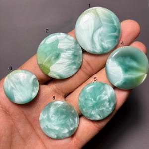 Pode incluir: Seis cabochões redondos e polidos em tons de verde-espuma e branco, cada um apresentando padrões rodopiantes semelhantes a nuvens. As pedras estão numeradas de um a seis, exibindo tamanhos variados e texturas marmoreadas únicas nas superfícies.