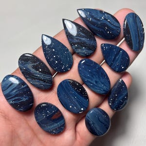 Könnte beinhalten: Eine Sammlung von elf polierten Cabochons in verschiedenen Oval- und Tropfenformen. Jedes Stück weist ein tief dunkelblaues und schwarzes Marmormuster mit verstreuten weißen Sprenkeln auf, die an einen Sternenhimmel erinnern, alle mit glatter Oberfläche.