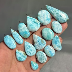 May include: Fifteen polished larimar gemstones in diverse shapes, including oval, teardrop, and elongated forms. The stones showcase a range of blue and white patterns, with some exhibiting a marbled appearance. The gemstones are numbered 1 to 15.