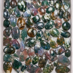 May include: A collection of polished moss agate cabochons in various shapes and sizes, including oval, round, and rectangular forms. Each stone features unique inclusions of green, brown, and white mineral patterns within a translucent base.