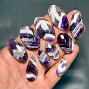 Può includere: Dieci cabochon di ametista chevron numerati da uno a dieci, caratterizzati da distinti motivi a bande a forma di V viola e bianchi. Queste gemme lisce, di forma ovale e a goccia, presentano una finitura lucida con variazioni di colore naturali.