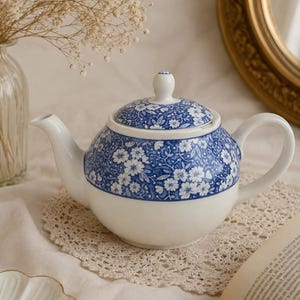 Puede incluir: Una tetera de cerámica blanca con un patrón floral azul en la mitad superior y en la tapa. La tetera tiene un asa curva, un pico vertedor y una tapa a juego con un pequeño pomo. El diseño consiste en flores y hojas blancas sobre fondo azul.
