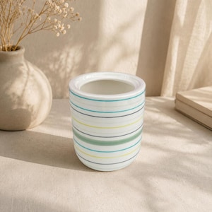 May include: A small, white ceramic container with horizontal stripes in shades of blue, green, and yellow. The container is cylindrical with a slightly flared top. A neutral background with a vase and dried flowers is visible.