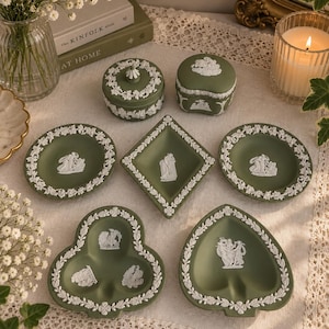 Puede incluir: Una colección de piezas de cerámica jasperware en color verde salvia con relieves neoclásicos blancos. El conjunto incluye dos cajas con tapa, dos platos redondos, un plato en forma de diamante, uno de trébol y otro en forma de pica.