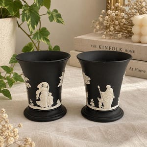 May include: A pair of matte black ceramic vases featuring white relief figures of classical scenes. Each vase has a flared rim, a tapered body, and a circular base. The white raised designs depict figures in flowing garments and trees.