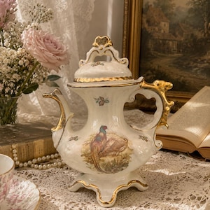 May include: A cream-coloured porcelain teapot with gold trim and a pheasant illustration. The teapot has a curved handle and a decorative lid. The design includes floral accents and is set on a lace tablecloth.