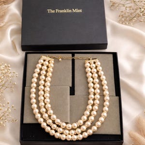 May include: A three-strand pearl necklace presented in a black box from The Franklin Mint. The necklace features cream-coloured pearls with small, sparkling accents between each strand. The box is open, revealing the necklace on a grey velvet insert.