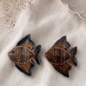 May include: Two ceramic fish figurines featuring a glossy brown body with textured scales and dark blue-black fins and tails. Each fish has a distinct eye and a sculpted shape, showcasing a smooth, glazed finish on the surface of the ceramic.