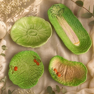 以下が含まれることがあります： 野菜の形をした4つのセラミック製サービング皿。皿は緑色で、レタス、セロリ、キャベツのデザインが特徴です。2つの皿には小さな赤いアクセントがあります。皿はニュートラルカラーの表面に配置されています。
