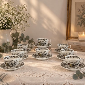 Può includere: Un set di sei tazze da tè in porcellana bianca con piattini abbinati, decorate con un dettagliato motivo floreale e di uccelli nei toni del marrone e del giallo. Le tazze presentano un manico curvo e un bordo largo, disposte su una tovaglia di pizzo.