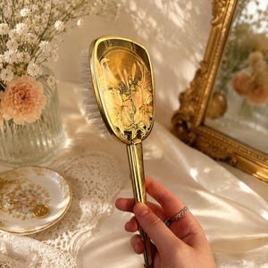 May include: A gold-toned hairbrush featuring a rectangular head with a decorative floral illustration of orange lilies against a dark background. The handle is slender and metallic, and the brush head includes white bristles for hair grooming.