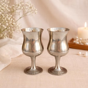May include: Two vintage silver goblets with a tulip-shaped bowl and a slender stem. The goblets are displayed on a light-colored surface, with a soft, neutral background. A lit candle and flowers are in the background.