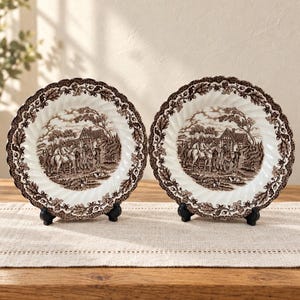 May include: Two round ceramic plates featuring a brown transferware pattern on a white background. The centre depicts a scene with horses and a building, while the scalloped rim is decorated with detailed oak leaves and acorns in brown.