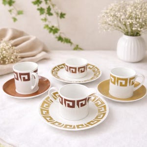 May include: A set of four vintage coffee cups and saucers. The white cups and saucers feature a geometric brown and gold design. The cups are on a white tablecloth with a vase of white flowers in the background.