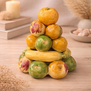 Può includere: Una scultura decorativa di frutta in ceramica con una piramide di arance, mele verdi e una banana gialla. La composizione è posta su una superficie di legno chiaro, con una candela accesa e piante secche sullo sfondo.