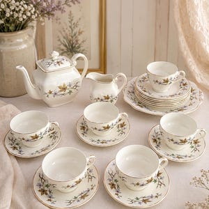 May include: A porcelain tea set comprising a teapot, a creamer, and six teacups with matching saucers. Each piece is off-white with a delicate botanical pattern of small blue flowers and golden-brown leaves, finished with a thin gold rim.